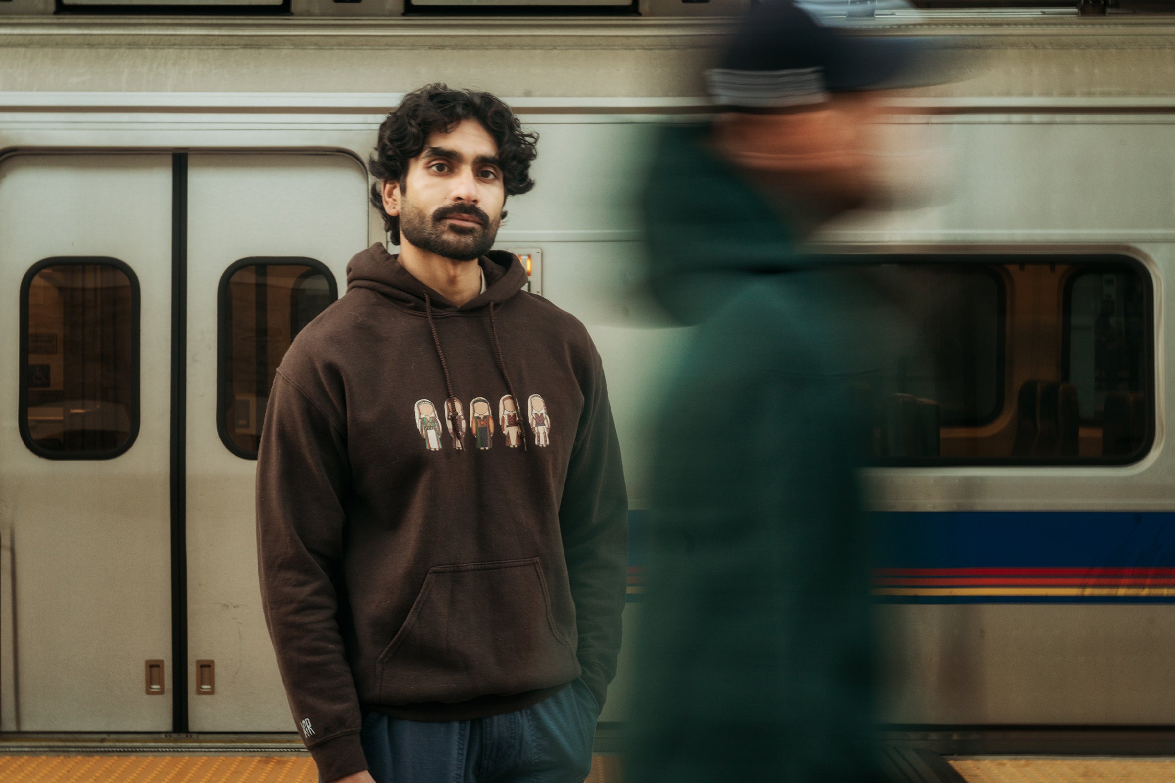 Man wearing a brown hoodie with a train in the background and blurry figure walking by
